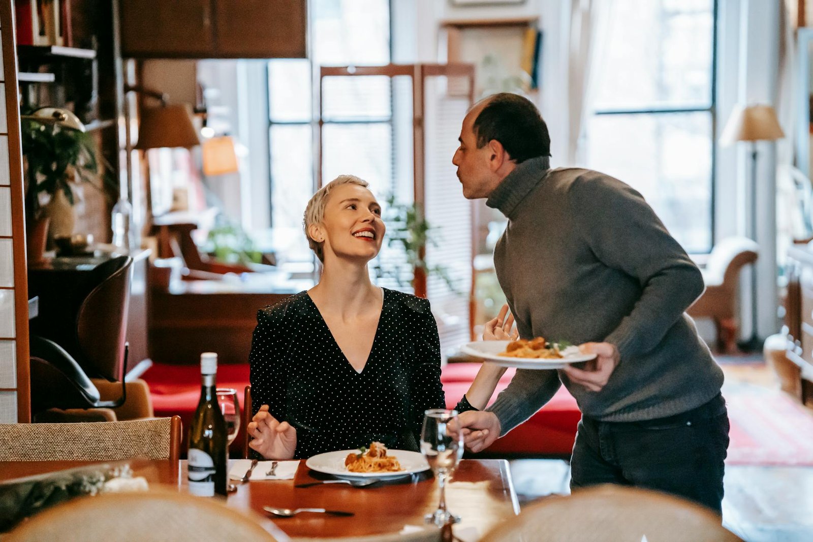 Adult couple having date in light room near table while male serving plates with pasta with meatballs near cutlery and bottle with wine near glasses while looking at each other
