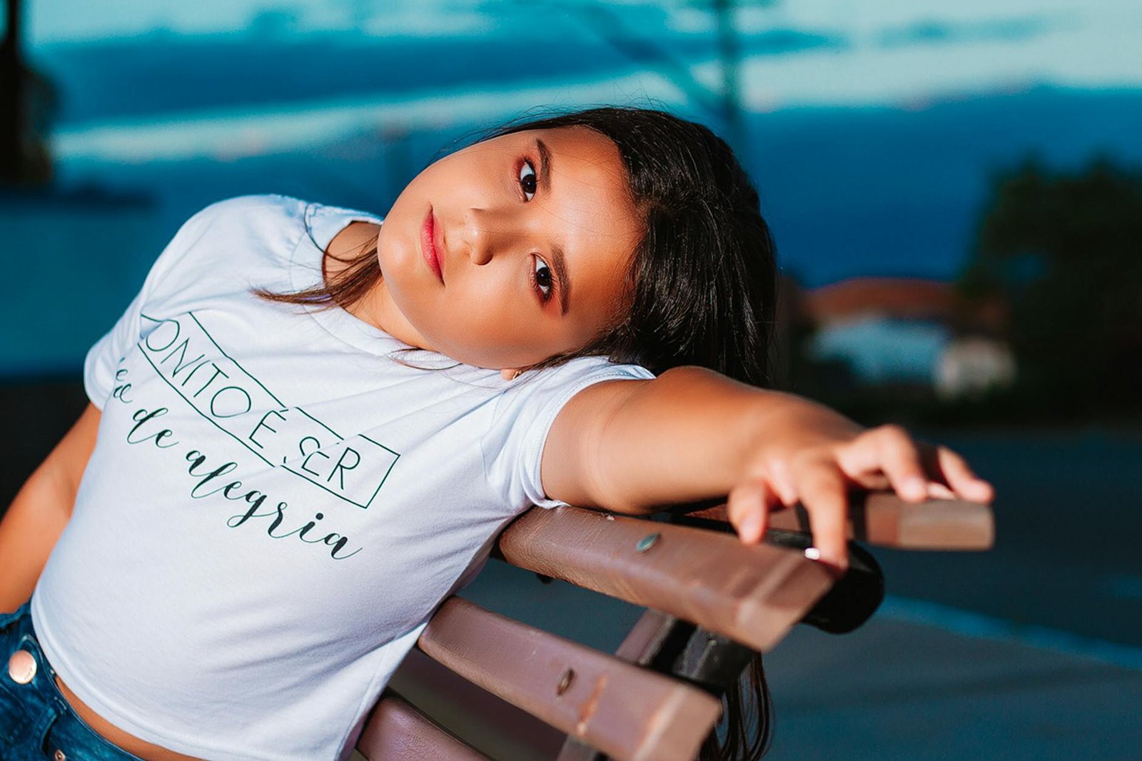 A young girl relaxing on a bench in a leisurely manner, wearing a white t-shirt with soft evening lighting.
