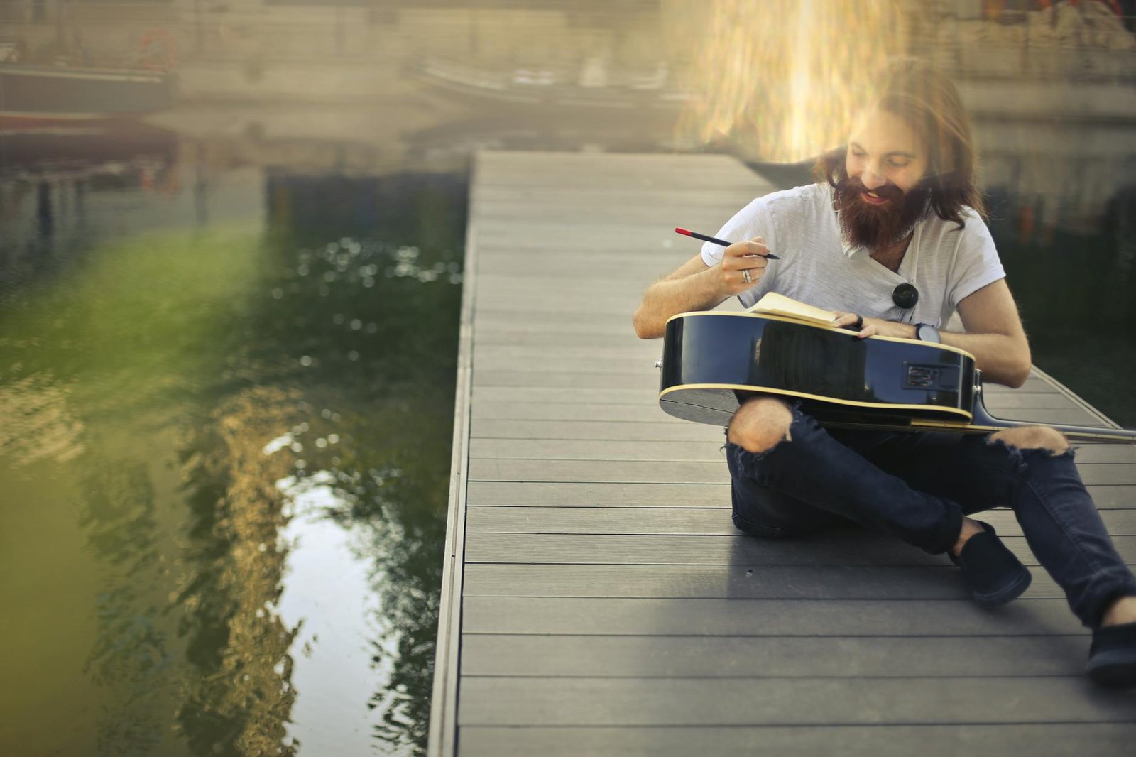 Smiling man on dock playing guitar and writing notes in a notebook with sunlight.