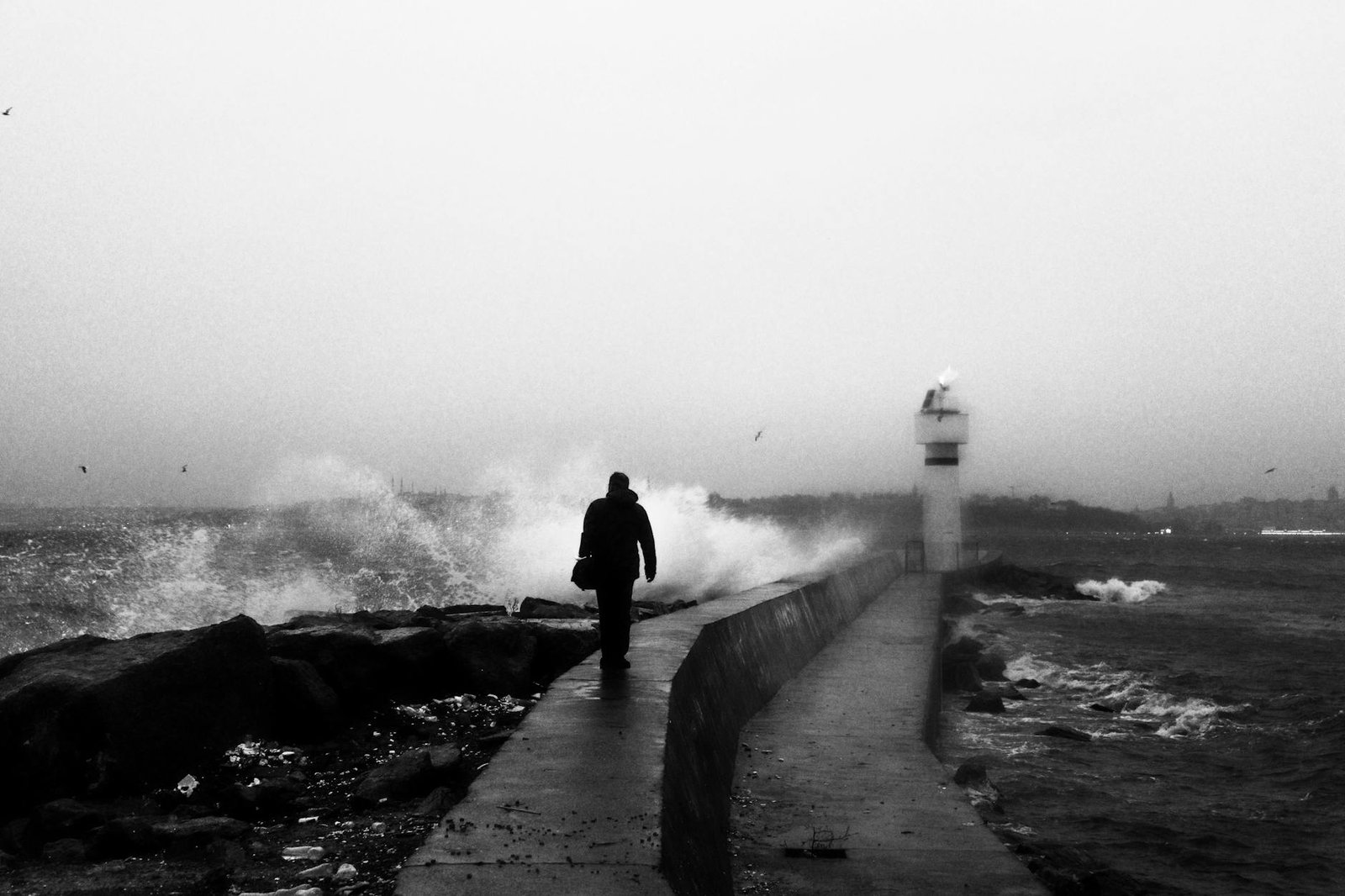 A lone silhouette walking on a breakwater with crashing waves and lighthouse in monochrome.