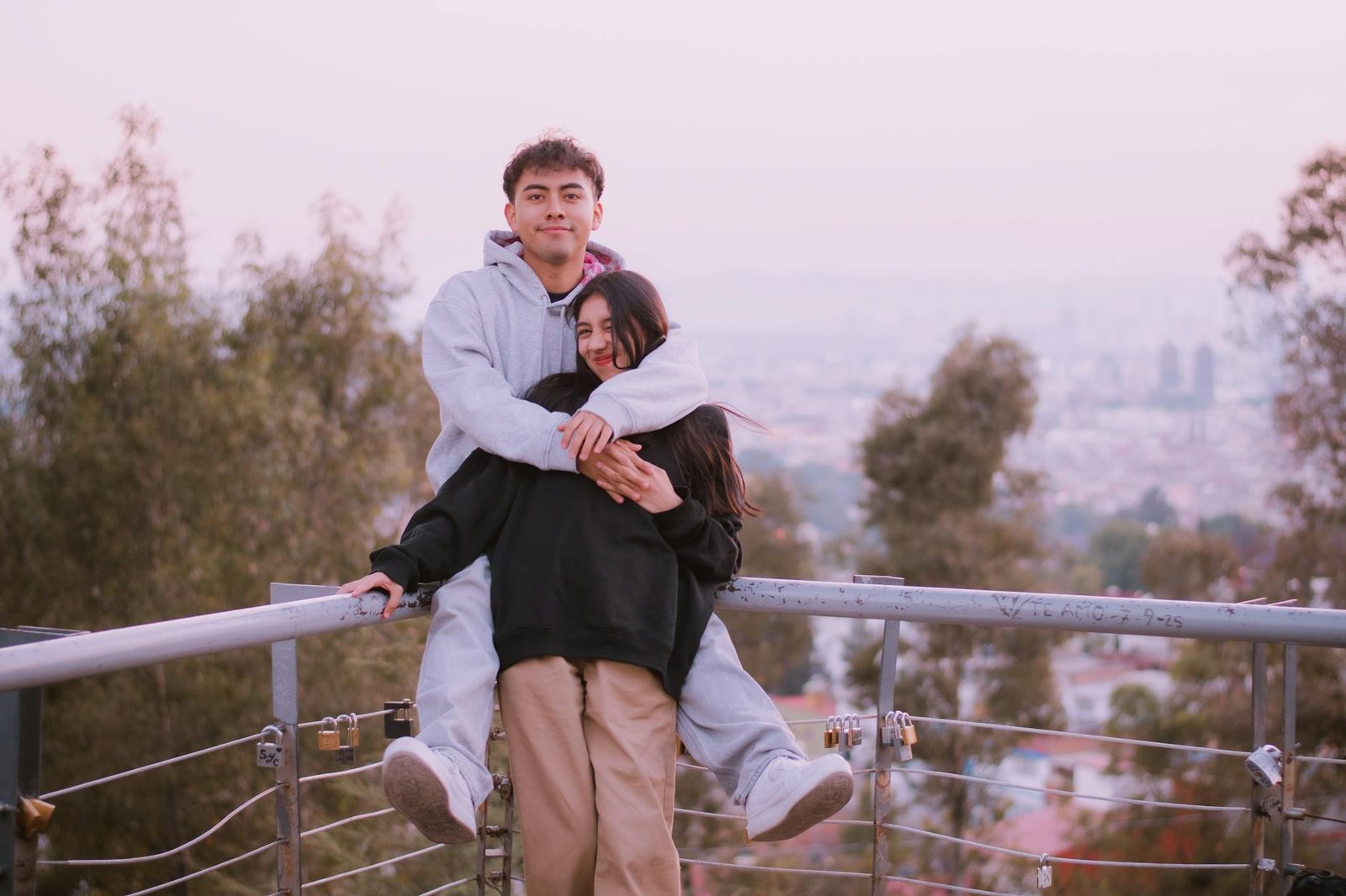 Young couple embraces on a railing overlooking a cityscape at dusk, surrounded by trees and soft evening light.