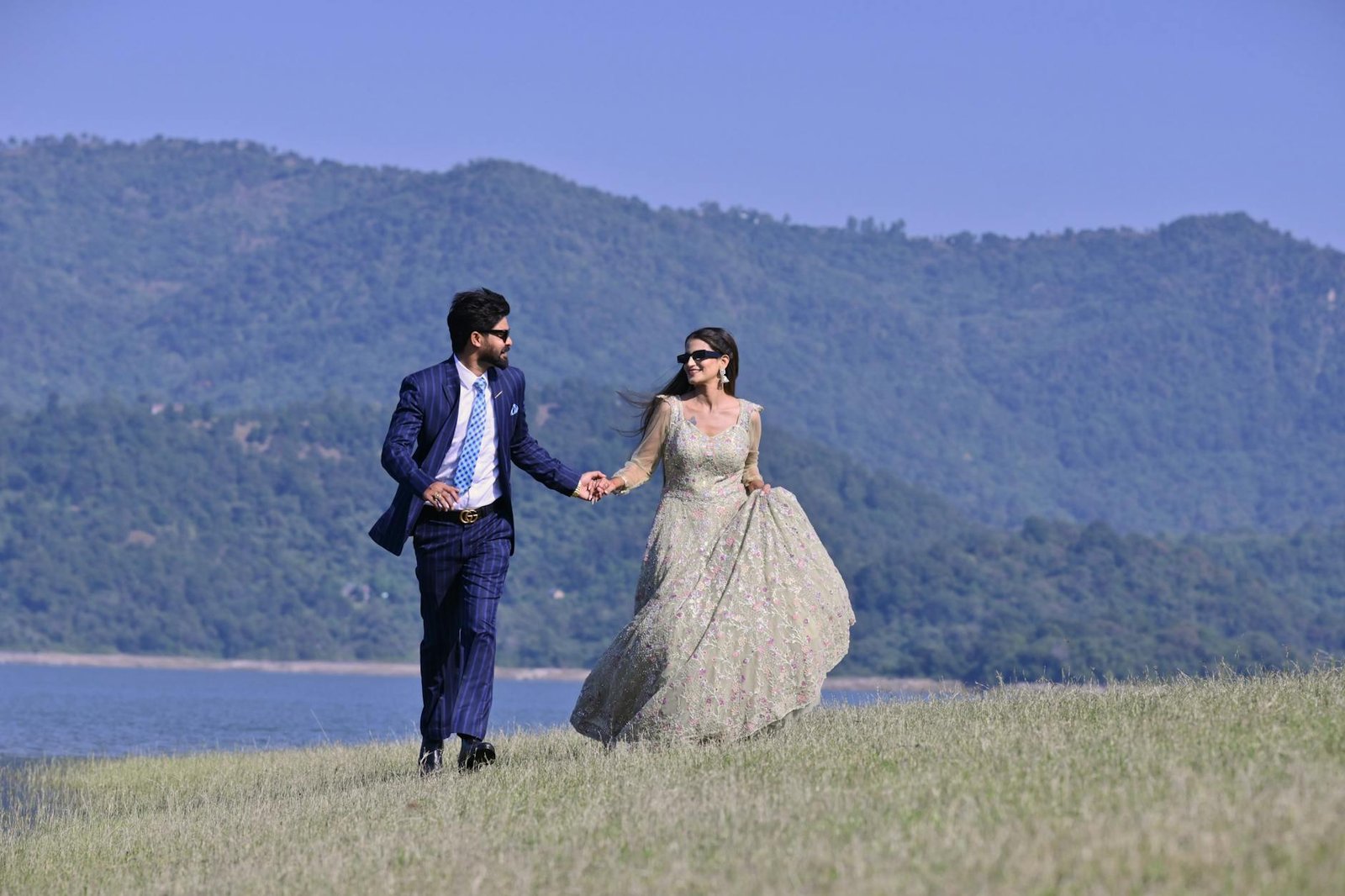 A couple in elegant wedding attire walking hand in hand by a picturesque riverbank in Una, India.
