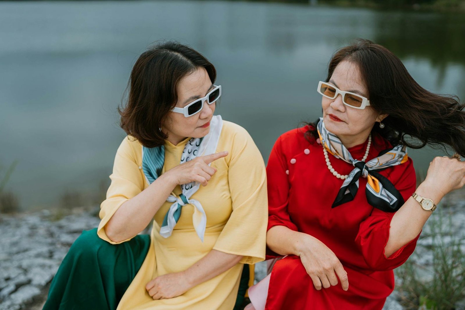 Two women in colorful ao dai sit by a riverside, exuding style and grace in a serene outdoor setting.