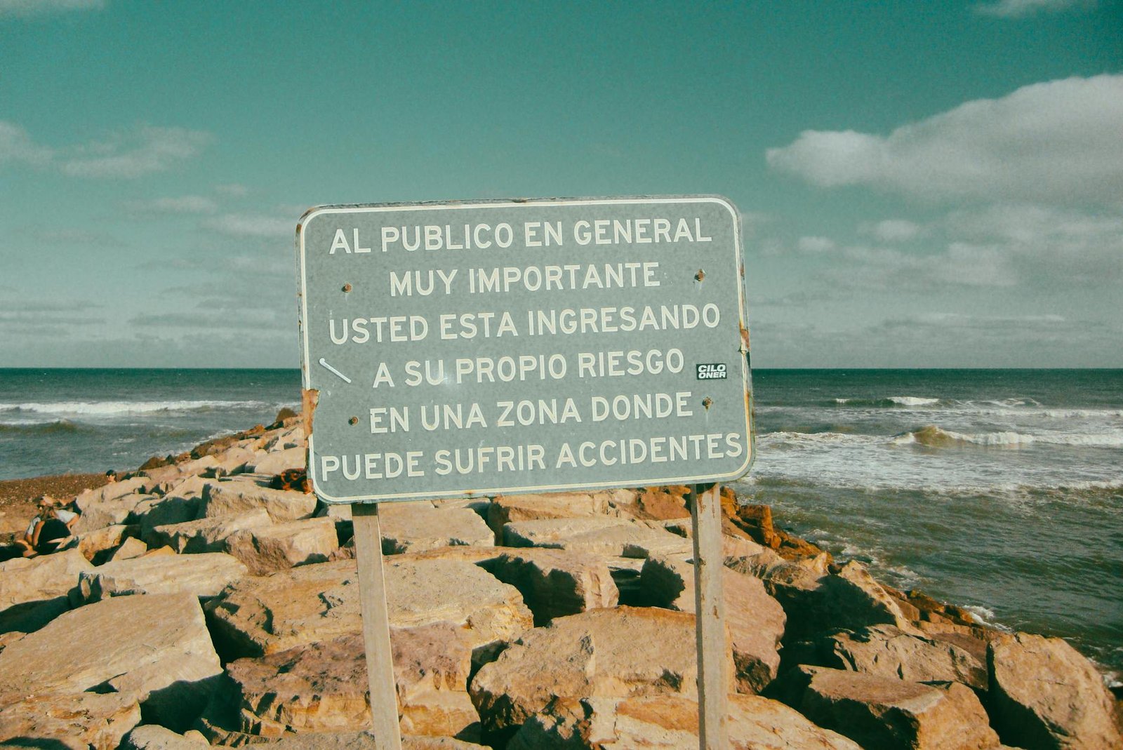 Spanish warning sign by rocky coast with crashing waves under clear skies.