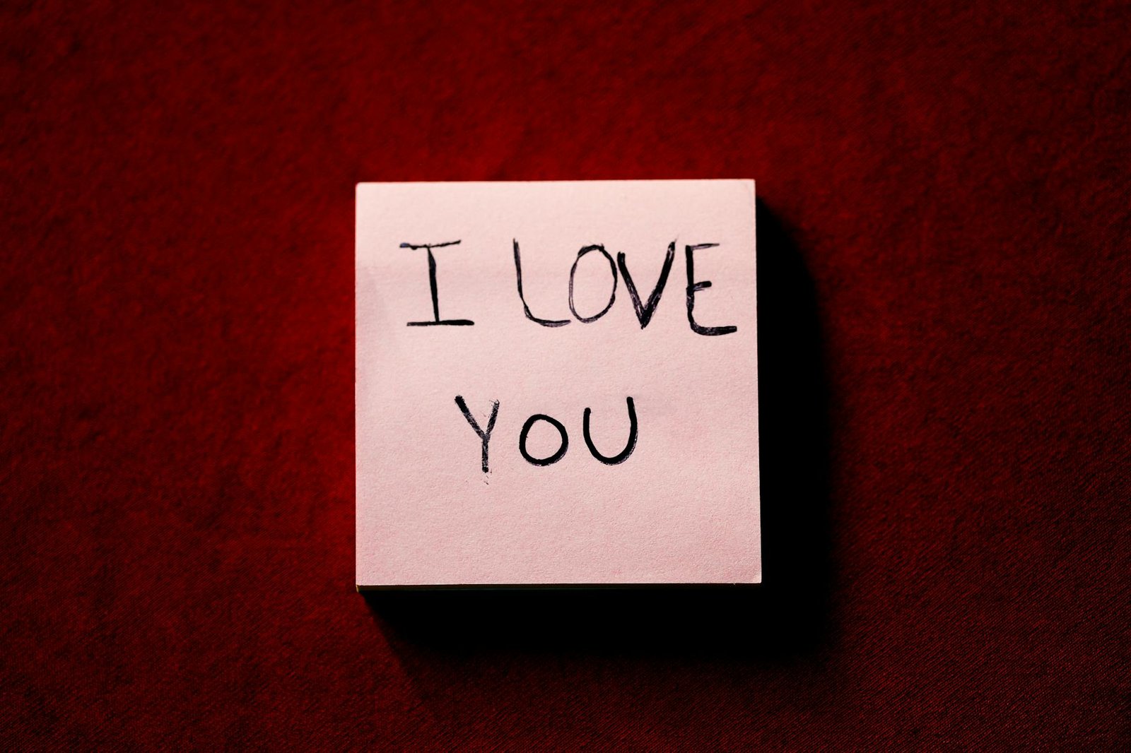 A close-up of a note with 'I Love You' written on it, placed on a red textured background.