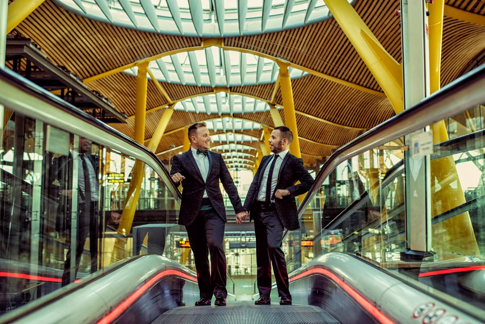 A joyful gay couple in suits holding hands on an escalator in a modern train station.
