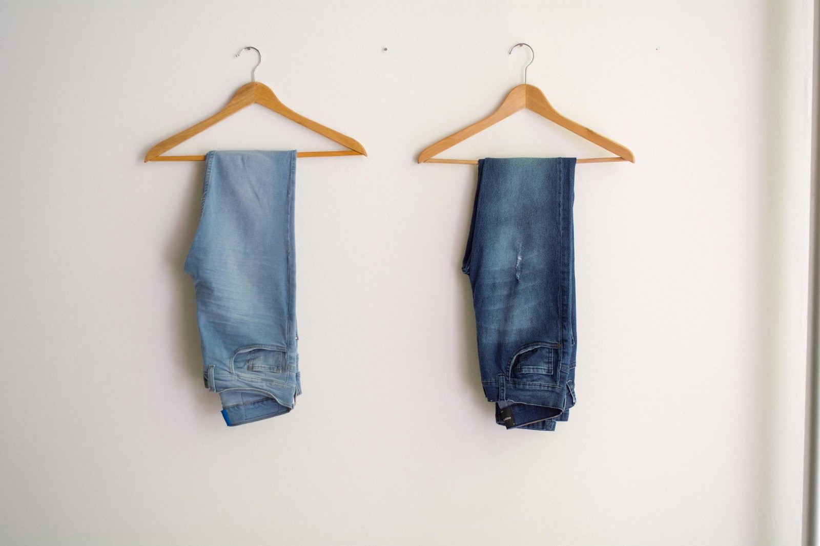 Two pairs of blue denim jeans neatly hanging on wooden hangers against a plain wall.